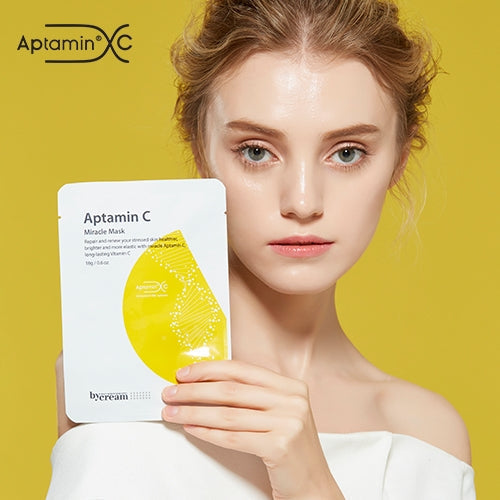 A person holding Aptamin C Miracle Mask package against a vibrant yellow background.